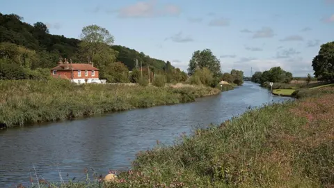 Getty Images A river curves through grass and wildflowers. A brick red and white house can be seen on the far bank. The sky is blue with some fluffy clouds.