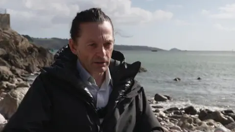 Dr Gavin Tilstone is in a black coat, black trousers and white shirt. He is sat on a rock on a rocky beach. He is facing the camera. The sea is in the background. The sky is blue with some white clouds.