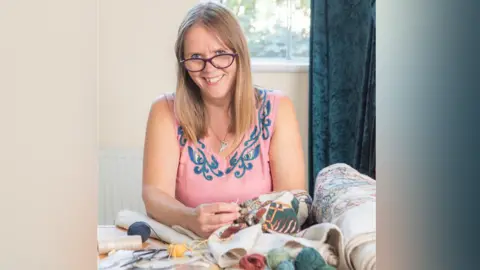 Search Press/Mark Davison Mia Hansson has blonde hair and glasses and is smiling at the camera, holding a needle and working on a colourful embroidery. She is wearing a pink sleeveless top decorated with a blue design.
