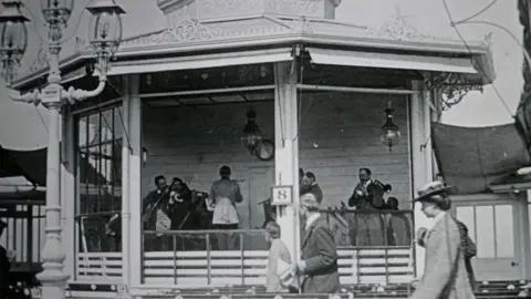Cromer Museum A black and white image of musicians playing in a bandstand, as three figures walk across the foreground