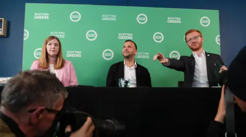 PA Media Gillian Mackay, Zack Polanski and Ross Greer sitting behind a table at a press conference, with a Scottish Greens backdrop behind them.