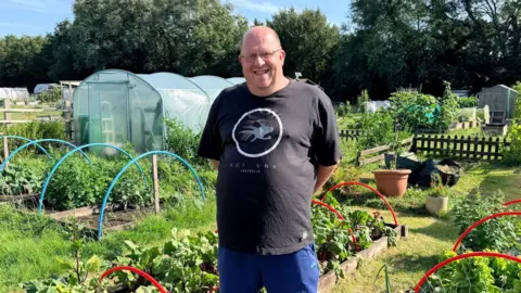 A man in a black tshirt and blue shorts smiles at the camera. Surrounding him are plots of crops with blue and red hoops over the planters.