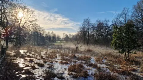 Askham Bog nature reserve photographed on a bright, clear morning. The ground is covered with a mix of patchy snow and frozen puddles, as well as brown grasses and low vegetation.