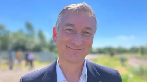 Simon Bachelor smiles and looks into the camera. He has grey hair. He's wearing a white shirt and a blue suit jacket.