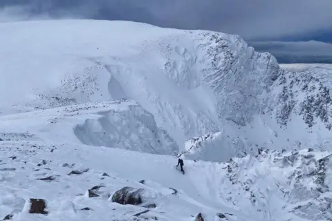 SAIS Northern Cairngorms A climber walks up a snow-covered slope in the Northern Cairngorms. He is dwarfed by a snowy mountain rising before him.