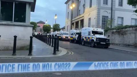 A number of police Land Rovers can be seen in a row near to Nailors Row in Londonderry. Black bollards line the other side of the road. A large pink building can be seen in the background as well.  