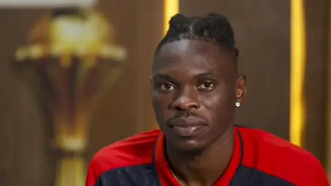 Sudan forward John Mano, visible from shoulders up in a red and black t-shirt, stares intently into the camera lens. A stud earring is visible in the lobe of his left ear. Blurred in the background over his left shoulder is a large replica of the golden Africa Cup of Nations trophy