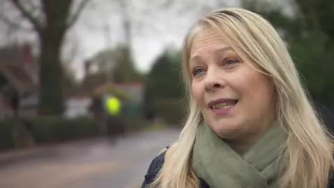 Hannah Colls has long blonde hair and is wearing a green scarf and blue coat. Behind her out of focus is a road.