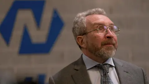 Wildcard Eddie Marsan has short grey curly hair and a grey beard and brown moustache is looking up. He is wearing a grey suit jacket, light coloured shirt and patterned tie. The background behind him is blurred.