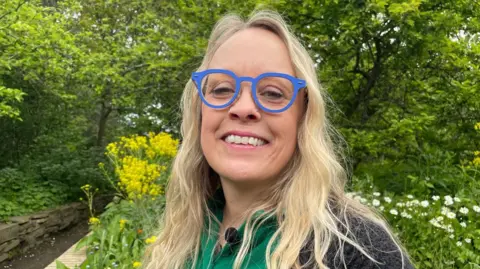 BBC/Emilia Martinez-Barnfather Karen Dobson, a middle-aged woman with blonde hair and bright blue glasses. She is, also, smiling. She is wearing a green polo shirt and black, knitted cardigan. Behind her are bright yellow flowers which have been planted in the garden, and beyond that, we can see bushes and trees rising out of frame.