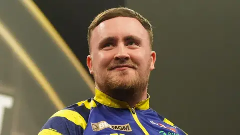 PA Media Darts player Luke Littler, in a blue and yellow top and smiling