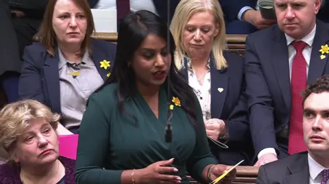 One MP talking (wearing a green jersey dress) and others listening in the House of Commons. Most are wearing small yellow heart pin badges.
