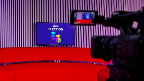 A red sofa in a broadcast studio. A TV screen is placed in the centre, displaying the words 'BBC Election 26' in white writing on a purple background. A TV camera is visible to the right of the image, out of focus.