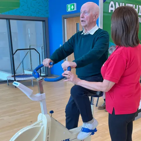 Joe Connolly on an exercise bike in a gym. He's sitting very upright and there is a medical professional beside him. Just her back is visible as she is talking to Joe. She has dark hair and a red t-shirt on, with black trousers.