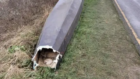 Phil Clarke The wreckage of a dragon boat, upside down, on the side of a country road