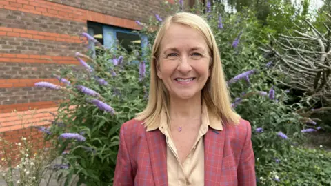 Leanne Rinne / BBC A woman, Katy Bourne, wearing a red suit jacket and cream top. She is stood in front of some flowers and smiling.