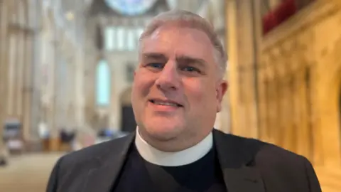 Canon Brown is wearing a black blazer and navy top. He has a clerical collar on. He is standing inside Lincoln Cathedral.