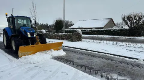 A snow plough is photographed on a snow-covered road, its yellow plough moving snow from the road.