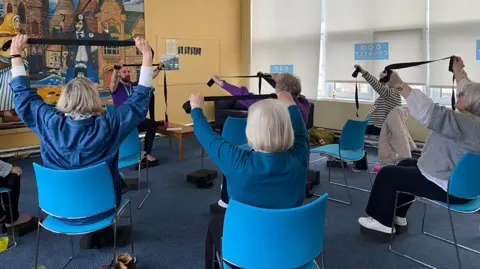 Five women sitting on blue chairs facing away from the camera. They are holding black yoga straps with both hands above their heads. A male instructor is facing the camera doing the same movement. 