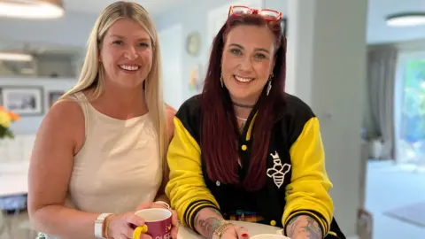 Emma Stirk and Dani Wallace smile for the camera and each hold a cup of tea 