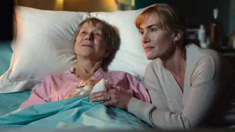 Kimberley French/Netflix Dame Helen Mirren in a hospital bed and Kate Winslet sitting beside her and holding her hand