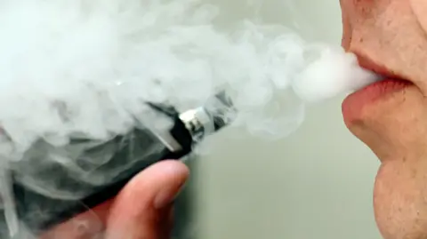 A close up of someone smoking a vape from a black device. Smoke is coming out of the smoker's mouth as they exhale.