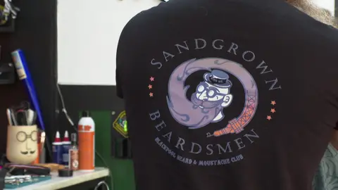 The back of a black T-shirt being worn by a man which reads Sandgrown Beardsmen - Blackpool Beard & Moustache Club