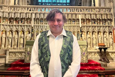 A man with brown hair stands inside a gothic minster church in front of the altar. He is wearing a white shirt and green patterned waistcoat. Behind him is a decorative screen containing rows of intricately carved stone statues.