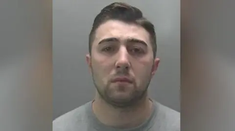 Custody image of Jordan Neale. He has short cropped dark hair and dark stubble, and is wearing a grey jumper.
