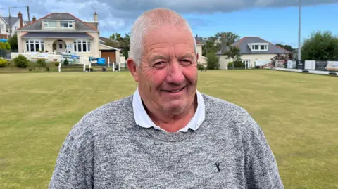 Richard Jones is stood in front of a large grassy field. He has short grey hair and is smiling and looking at the camera. 