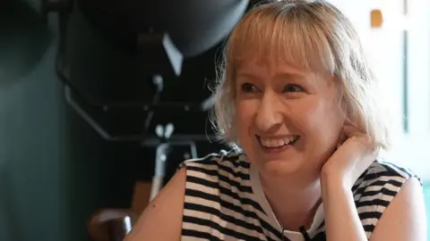 Laura Price has short blonde hair. She is wearing a blue and white striped vest top.