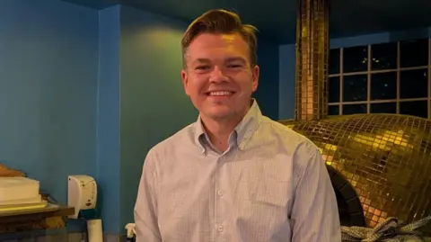 A wide shot of Adam smiling. He has short brown hair and is wearing a white shirt.