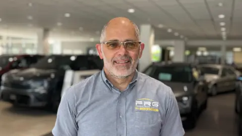 Robert Cornelius standing in a car show room. He is bald with a grey beard and is wearing tinted glasses. He is wearing a grey button up shirt with the Forest Road Garage logo on it.
