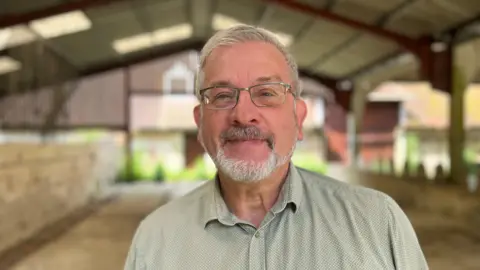 Guy Hall who has grey hair, beard and glasses standing in an old barn in Stanmer Park which he hopes his charity can use as a workshop and museum