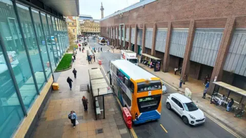 Newcastle City Council An overhead view of Blackett Street. A grey tarmac road, with a lane going each way, cuts through the city. The road is dotted with buses, a zebra crossing and other vehicles. There is a wide brown, tiled pavement on either side with four bus stop shelters and people wating in them. The street is then bordered by tall buildings. There is a line of windows on the left, and the grey striped design on the red-brick building on the right.