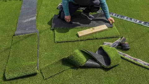 Miami Herald/Getty Images Sheets of artificial grass in a garden. There are off-cuts on top of them as well as a person, whose face cannot be seen, crouching down cutting a piece of the plastic grass. 