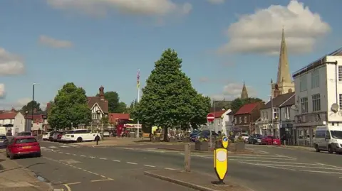 A town square surrounded by shops. Several cars are parked in the centre of the area which is dominated by two large trees. The spire of the local church is visible in the background set against a blue sky.
