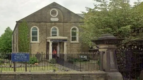 Providence Chapel on Huddersfield Road in Elland, West Yorkshire. The chapel building is surrounded by iron gates and a stone wall. There is a small lawned area near the entrance to the building. 