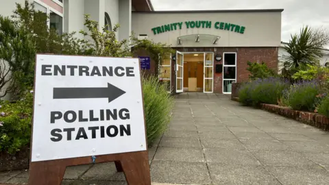 Close-up of a white sign with black writing on it pointing people towards the entrance of a polling station inside the Trinity Youth Centre in Jersey.