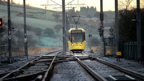 TfGM A yellow tram on a rural track. Foggy hills can be seen behind