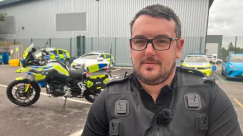 Roads Policing Inspector Matt Boiles from Avon and Somerset Police stands in a car park with police cars and motorbike behind him.