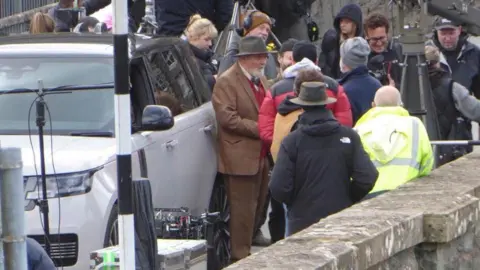 Nia Wyn Evans Sir Anthony Hopkins is stood by a Range Rover wearing a brown suit jacket and trousers, and a hat. He is surrounded by people from a production company for his new film.