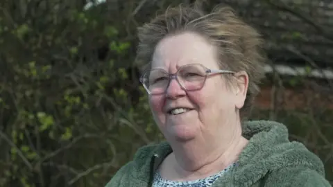 Gwen Olson, who is wearing a blue patterned top with a green fluffy jacket and glasses, is standing in a field as she is being interviewed. 