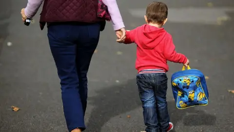 Generic image of a woman holding the hand of a small boy who is wearing jeans and a red hoodie and is holding a blue minions rucksack. They are walking away from the camera so their faces cannot be seen. The woman can be seen from the waist down and is wearing jeans and a dark red gillet.