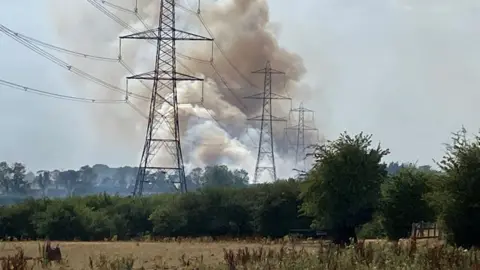 A rural landscape with a plume of smoke rising in the distance. Pic taken from the edge of a field, looking towards a line of trees and vegetation. Several large electricity transmission pylons dominate the scene, carrying multiple high-voltage power lines across the frame. Behind the tree line, a large column of grey and brown smoke is rising into the sky.