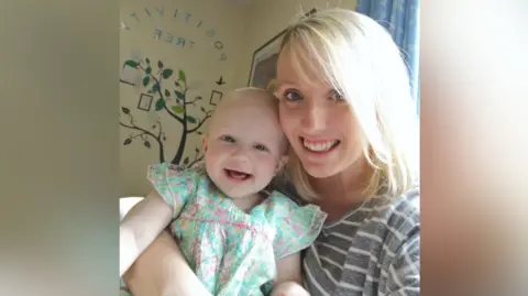 Handout Kaylee Avery, with blonde hair and a striped top, holds a baby in a flower-print dress inside the hospital ward.