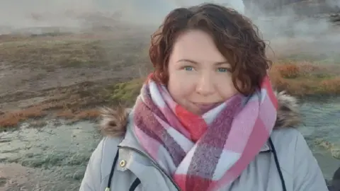 Emma Drinkwater Emma Drinkwater wearing a pink and white checked scarf and a grey coat. She has brown curly hair and is smiling at the camera. She has blue eyes.