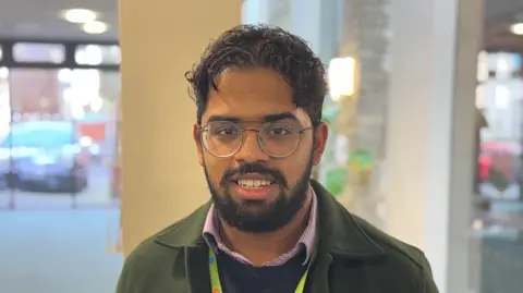 George King/BBC A man with dark hair and wearing glasses and a grey jacket smiling into the camera. 