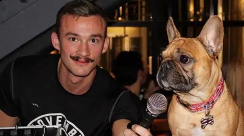 Fergus Crawley Fergus Crawley is wearing a black T-shirt and holding a microphone to the mouth of his pet bulldog sitting next to him. He is wearing a fake moustache.