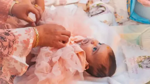 Foto keluarga Dalam foto ini bayi Sophia terlihat sedang berbaring mengenakan gaun ruffled pink lembut dan sedang menghisap dot. Dia memiliki rambut coklat jambul dan mata gelap yang besar.